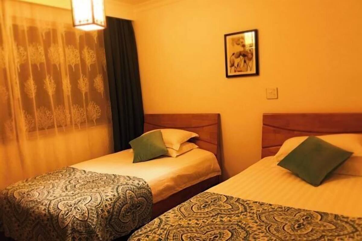 Een hotelkamer met twee bedden, een nachtkastje, een lamp, een ingelijst schilderij aan de muur en een raam met dunne gordijnen.