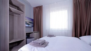 Une chambre d’hôtel avec un lit, un téléviseur fixé à un meuble en bois, une fenêtre avec des rideaux légers et un tableau au mur.