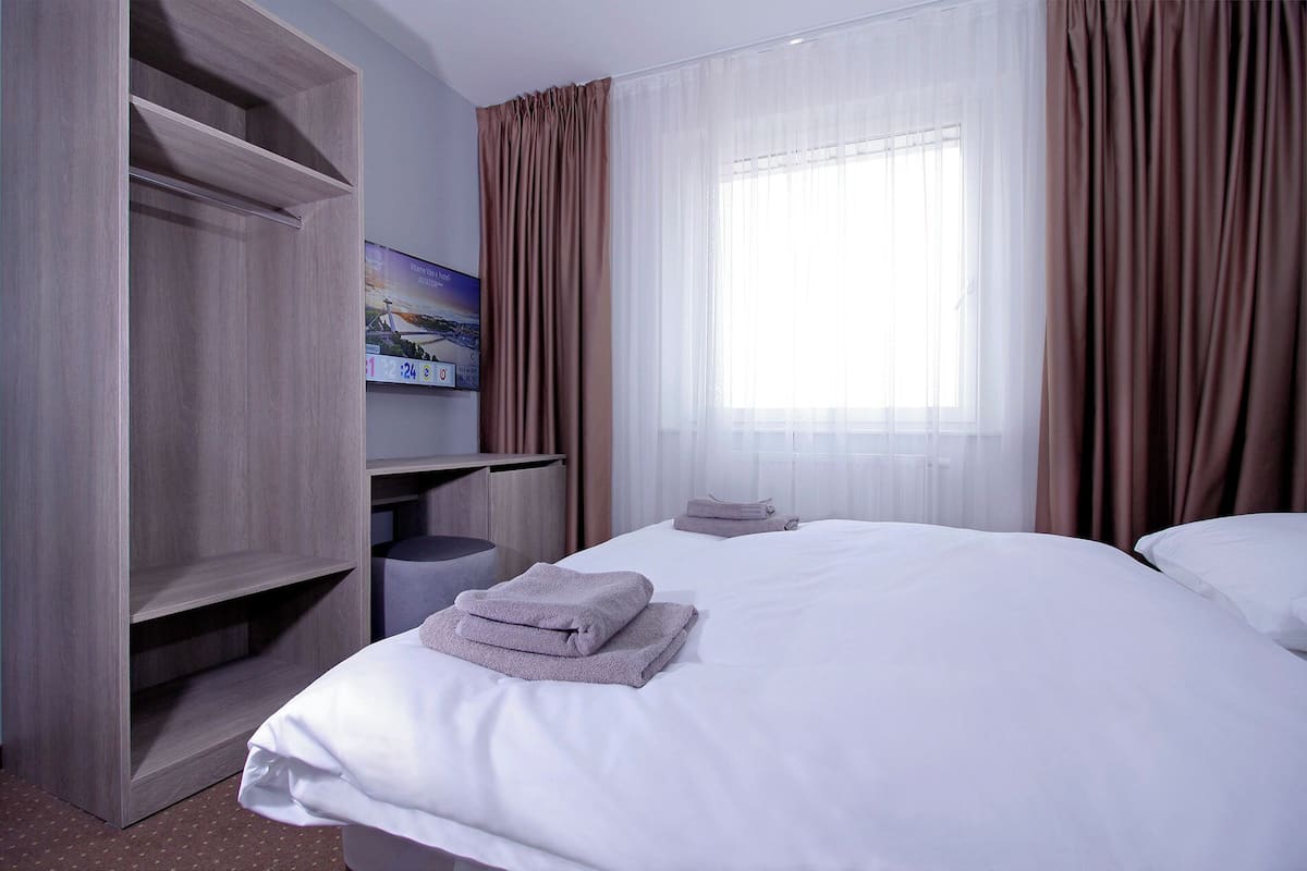 A hotel room with a bed, a TV mounted on a wooden cabinet, a window with sheer curtains, and a painting on the wall.