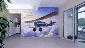 Un interior moderno con un mural de un avión volando entre nubes, una puerta de vidrio que da al exterior y una planta en maceta a la izquierda.