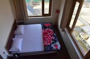 Una cama pequeña con ropa blanca y una manta estampada con motivos florales, una ventana con cortinas y vistas a un espacio exterior con rocas y plantas.