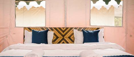 A bed with a patterned headboard, two windows with curtains, and rolled towels on the bed.