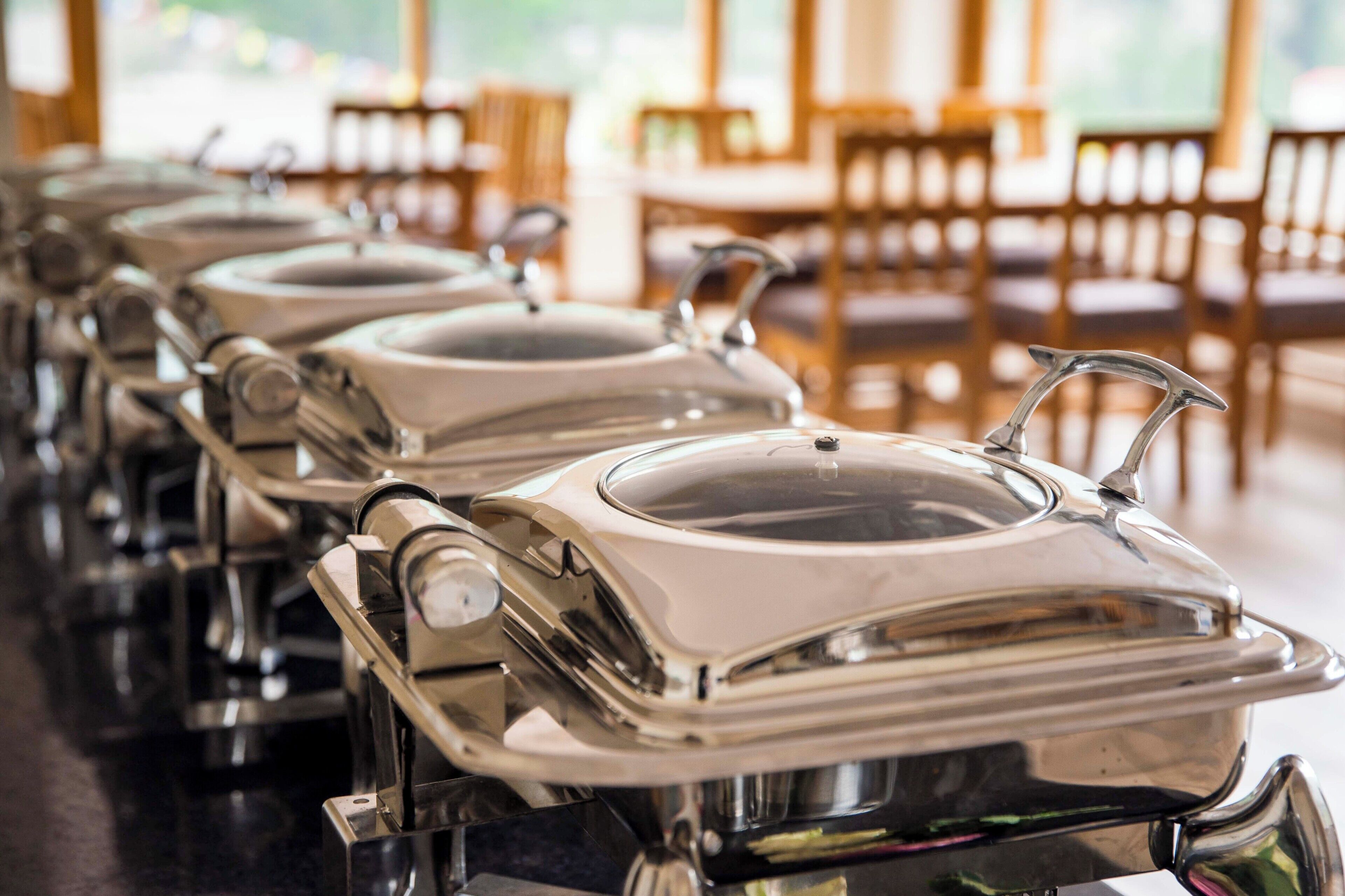 Une rangée de chariots de buffet en acier inoxydable, dotés de couvercles, est disposée dans un espace de restauration agrémenté de chaises et de tables en bois.