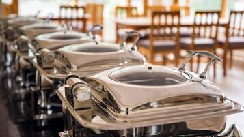 Une rangée de chariots de buffet en acier inoxydable, dotés de couvercles, est disposée dans un espace de restauration agrémenté de chaises et de tables en bois.