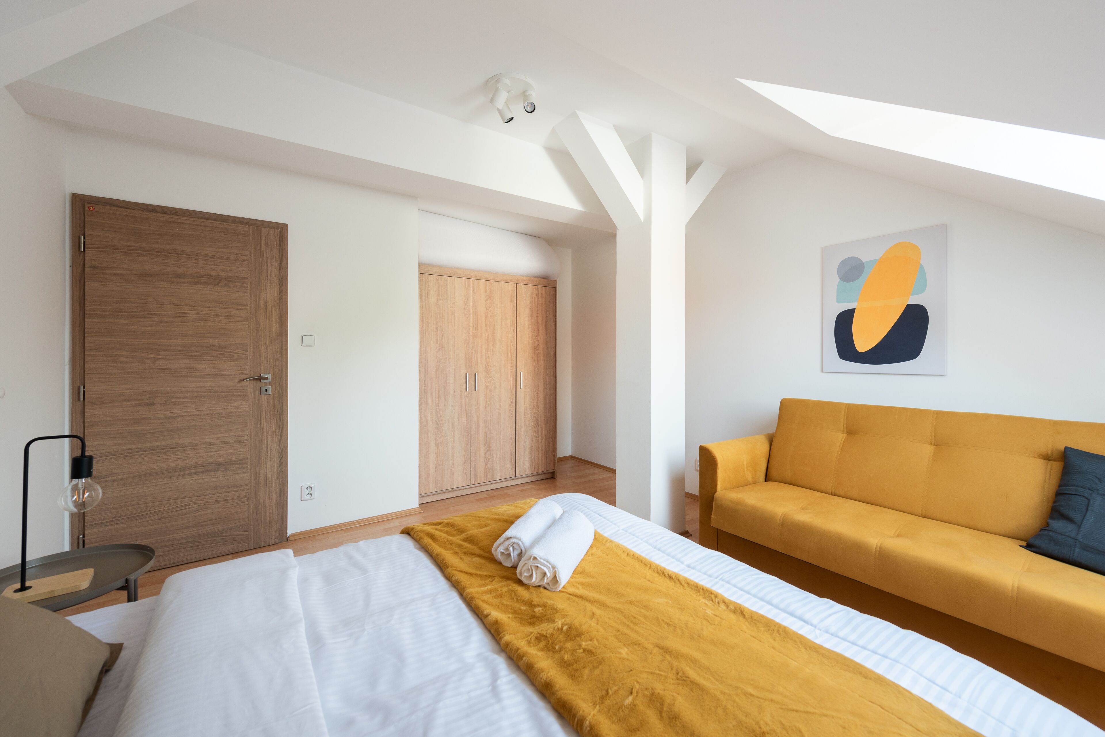 A modern hotel room with a bed, a sofa, a wooden wardrobe, and a painting on the wall.