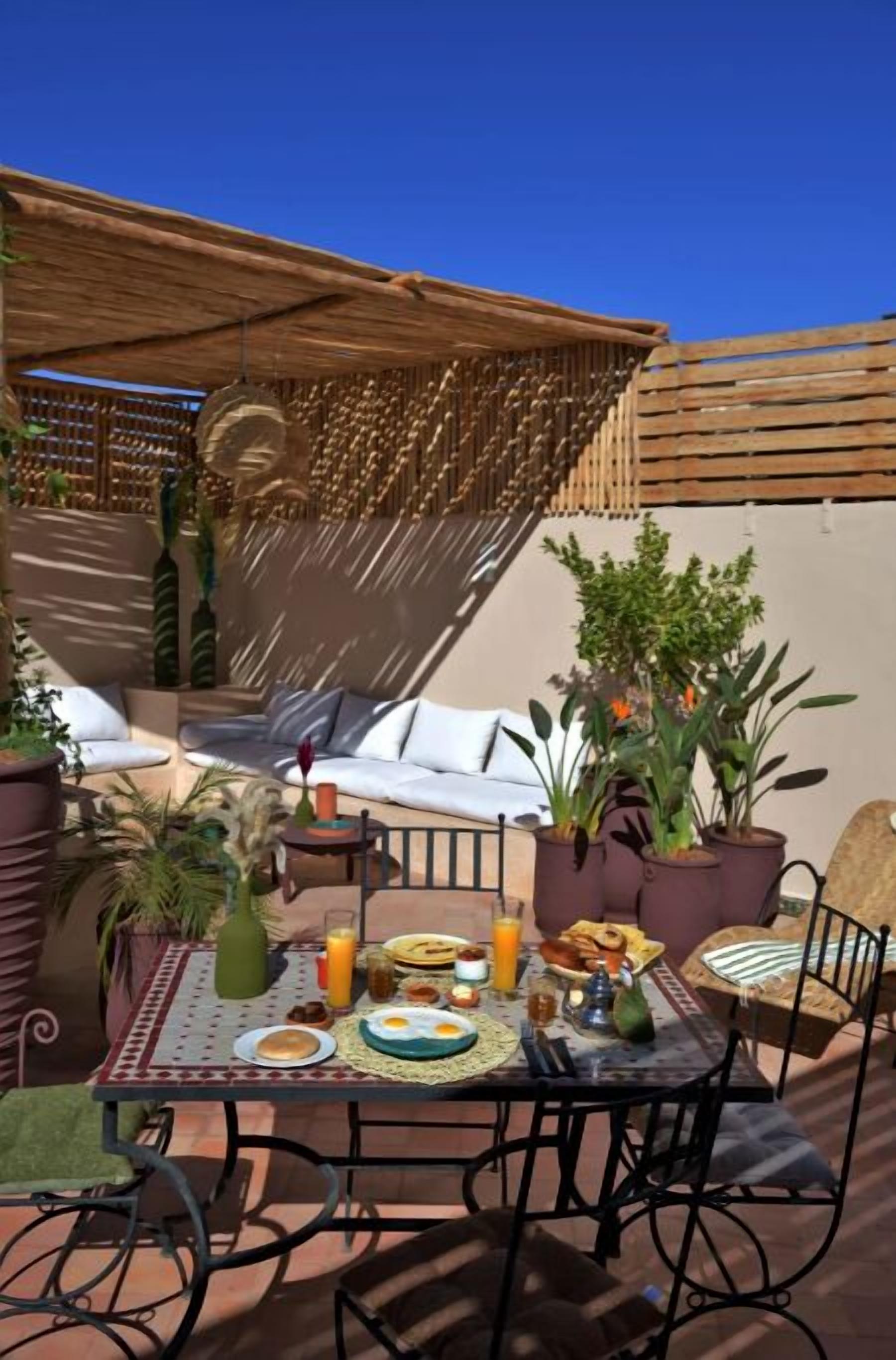 An outdoor dining area with a table set for two, surrounded by potted plants and a wooden pergola.