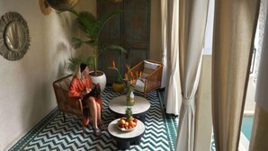 Ein Zimmer mit einem gemusterten Boden, einem runden Tisch mit Früchten, einer Couch, einem Sessel, einer Pendelleuchte und einem dekorativen Spiegel.