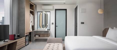 A modern hotel room with a large bed, a sitting area, a vanity with a mirror, and a view of the city.