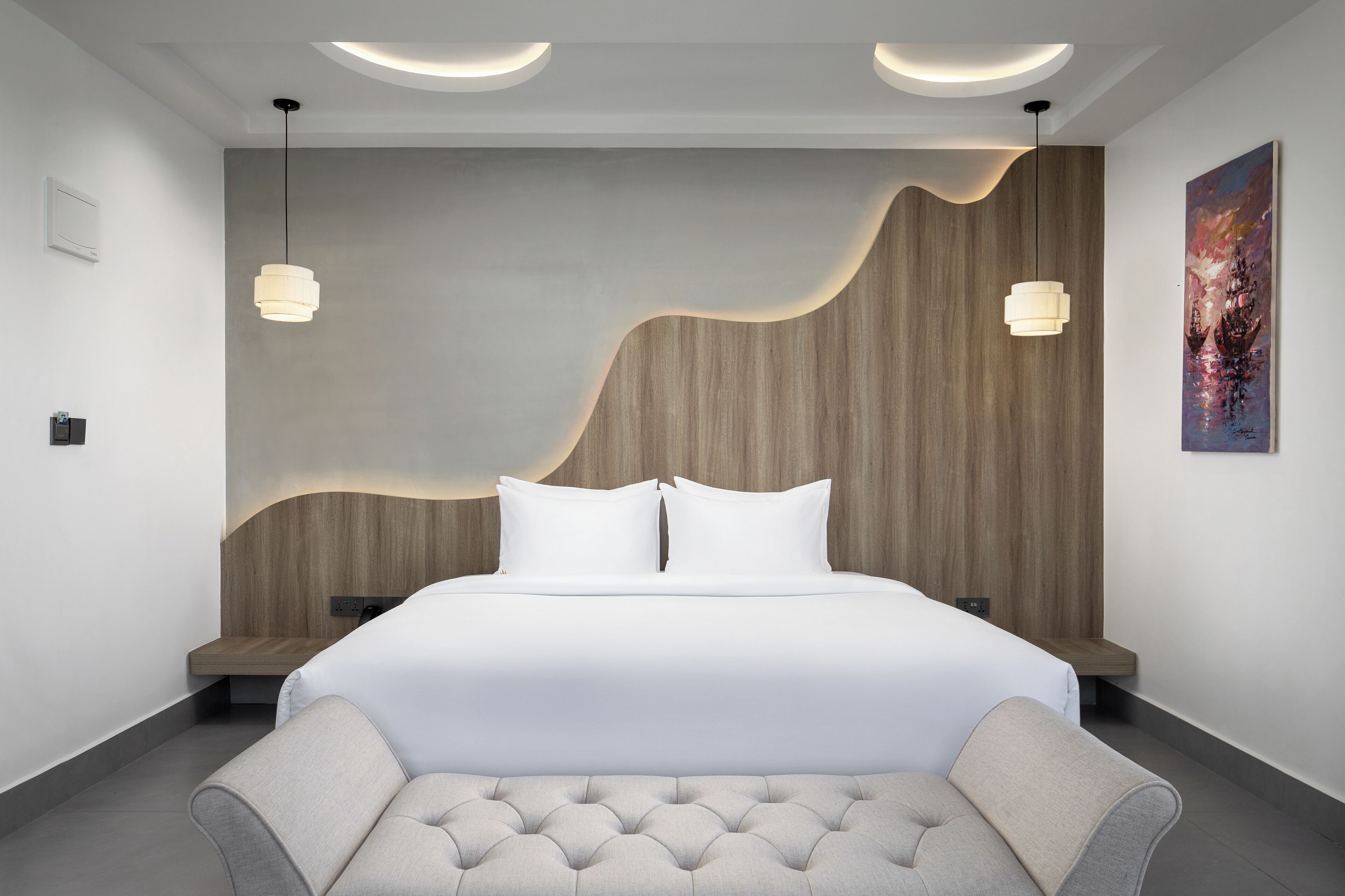 A modern bedroom with a large bed, two pendant lights, a wooden headboard, and a painting on the wall.