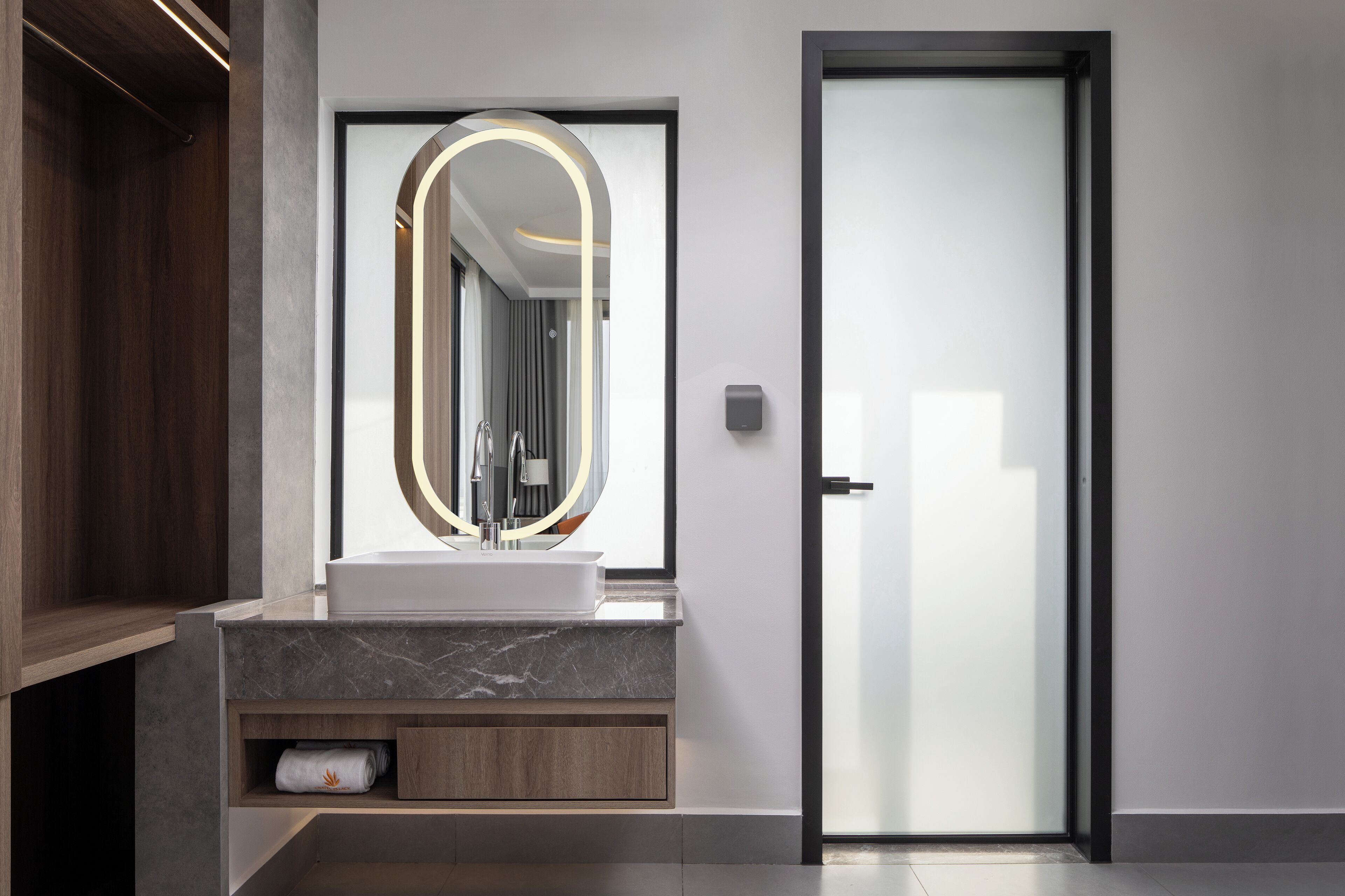 A modern bathroom with a marble sink, a large mirror with a circular frame, and a frosted glass door.