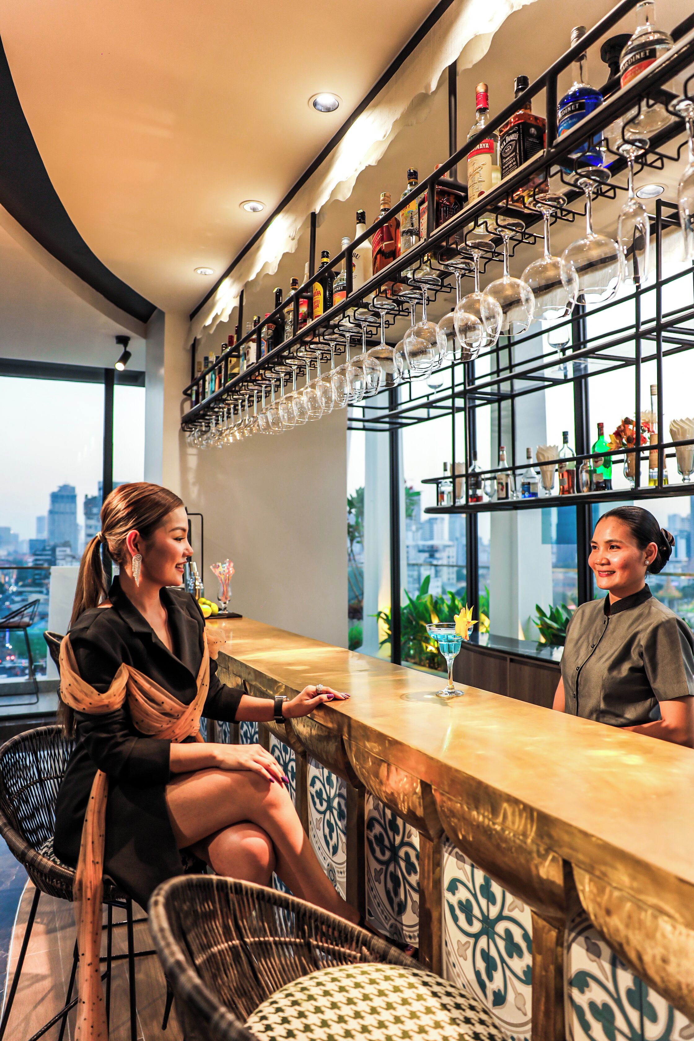 A bar area with a city view, wicker chairs, and a variety of bottles and glasses on shelves.