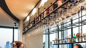 A bar area with a city view, wicker chairs, and a variety of bottles and glasses on shelves.