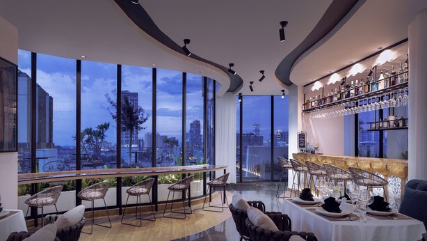 Un restaurante moderno con vistas panorámicas de la ciudad, una zona de bar con estantes llenos de botellas, y una combinación de sillas de mimbre y tapizadas.