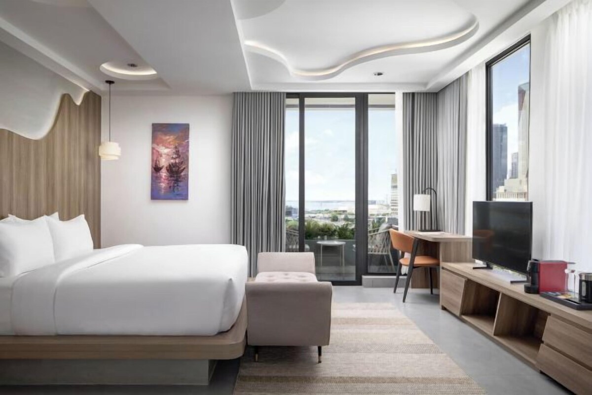 Habitación de hotel moderna con una cama grande, un escritorio y una televisión. Se observa una vista de la ciudad a través de amplios ventanales.
