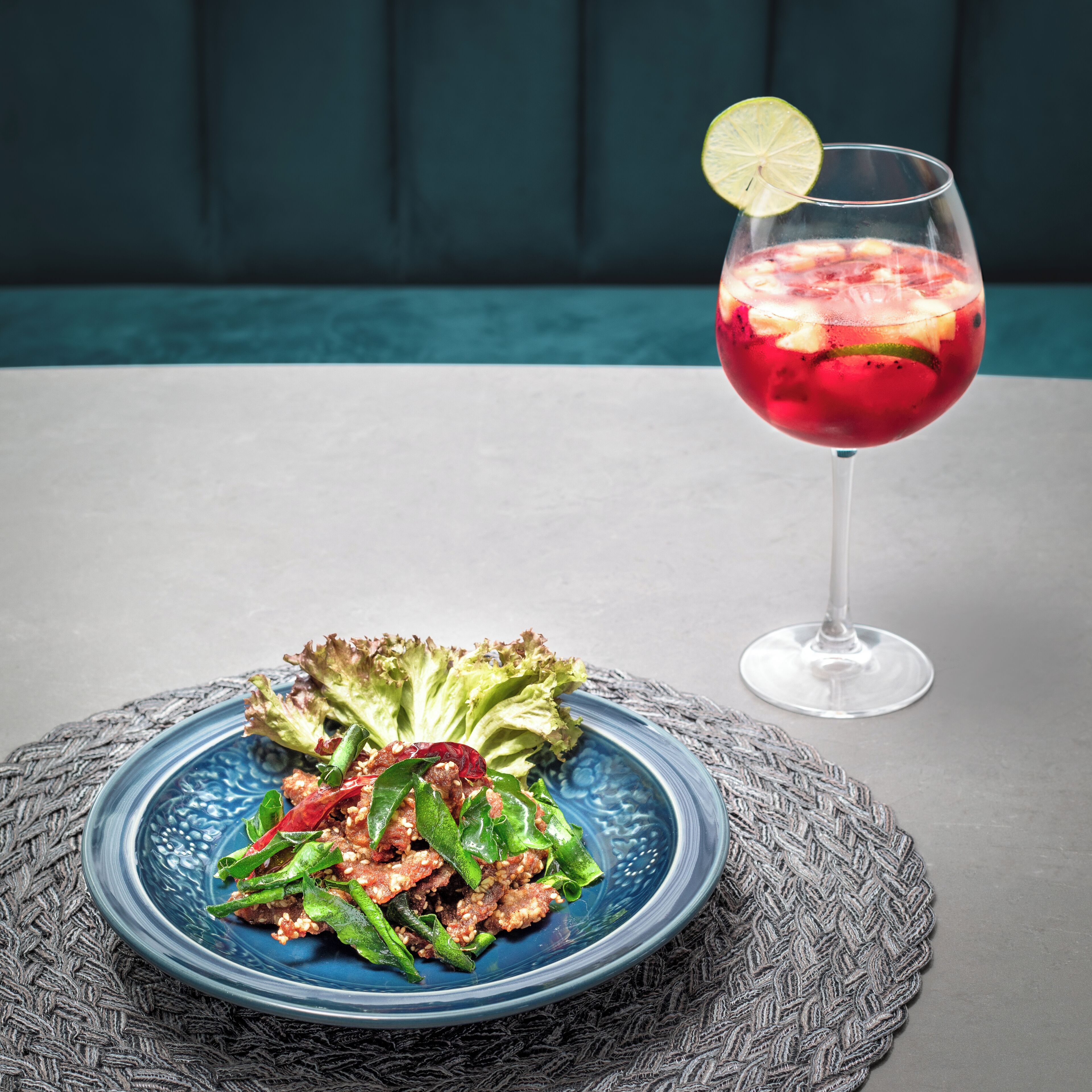 Una mesa con un plato azul de ensalada y un vaso de bebida roja adornado con lima.