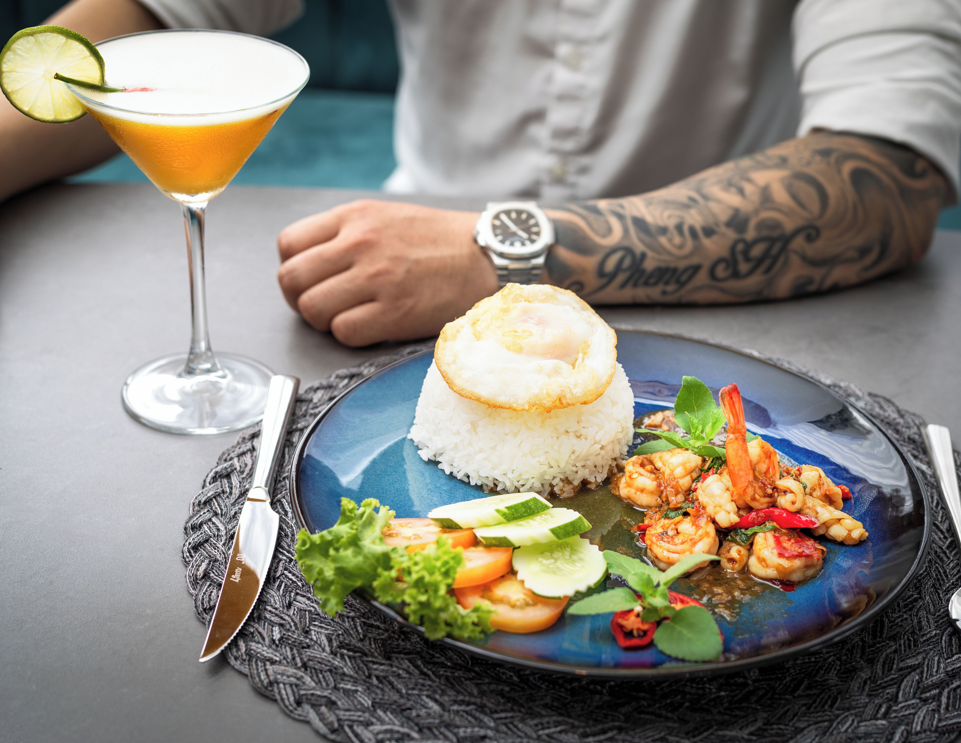 Lautasella on valkoista riisiä, jonka päällä on paistettu kananmuna, katkarapuja ja kasviksia. Annoksen kanssa tarjoillaan cocktail, joka on koristeltu limenviipaleella.