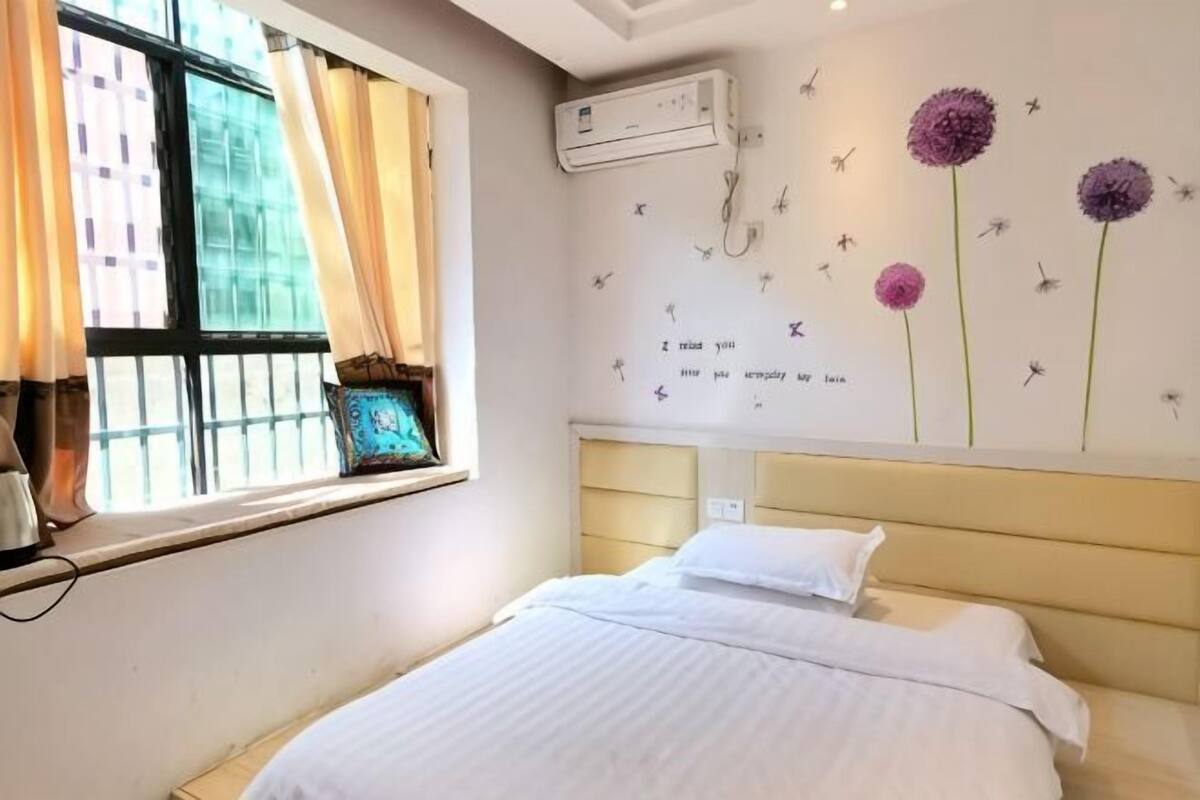 Een slaapkamer met een bed, een raam met gordijnen, een wandairconditioner en decoratieve wandkunst.
