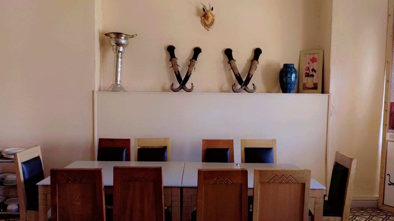 A dining area with a long table and six chairs, a decorative wall with a skull and two large hooks, and a blue vase.