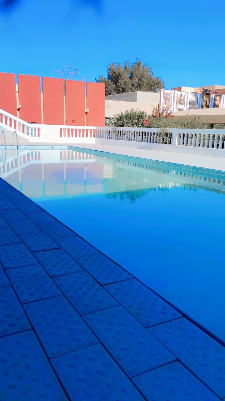 A swimming pool with a tiled deck, surrounded by a white fence and a red building in the background.