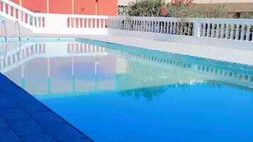 A swimming pool with a tiled deck, surrounded by a white fence and a red building in the background.
