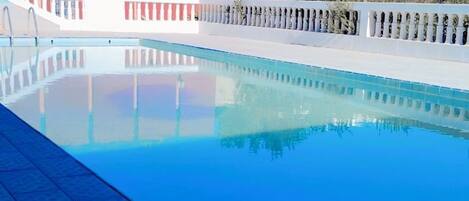 A swimming pool with a tiled deck, surrounded by a white fence and a red building in the background.