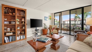 Ein geräumiges Wohnzimmer mit einer Holzbücherwand, einem Flachbildfernseher, einem gemütlichen Sofa und einem Balkon mit Blick auf Palmen.