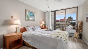 Ein Schlafzimmer mit Bett, Nachttischen, Deckenventilator, großem Fenster mit Vorhängen und Blick ins Freie.