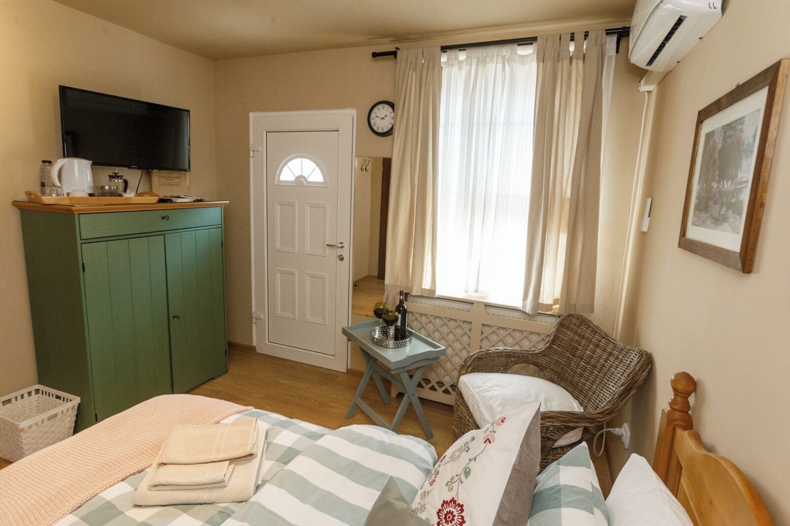 A cozy room with a bed, a green cabinet, a small table with a bottle, a wicker chair, and a window with curtains.