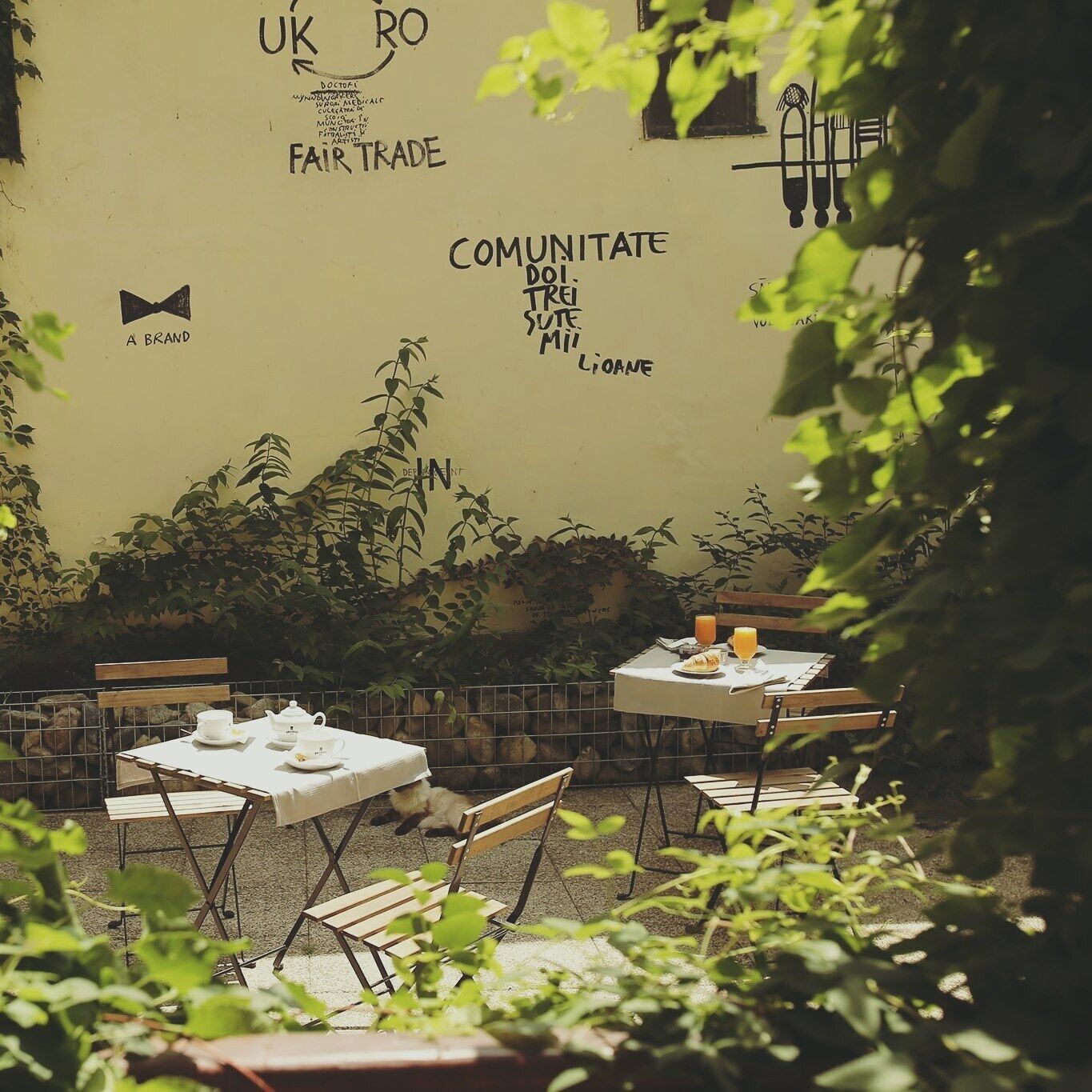 Een terras met tafels en stoelen, omgeven door groen en een muur met diverse teksten en logo's.