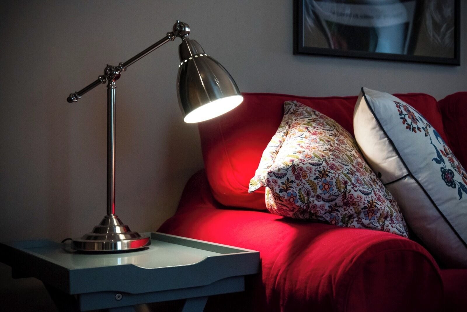 A desk lamp on a bedside table next to a red sofa with floral cushions.