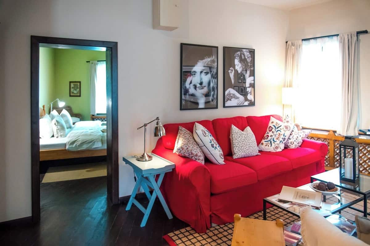 A red sofa with patterned cushions, a small table with a lamp, and a mirror reflecting a bed with pillows.