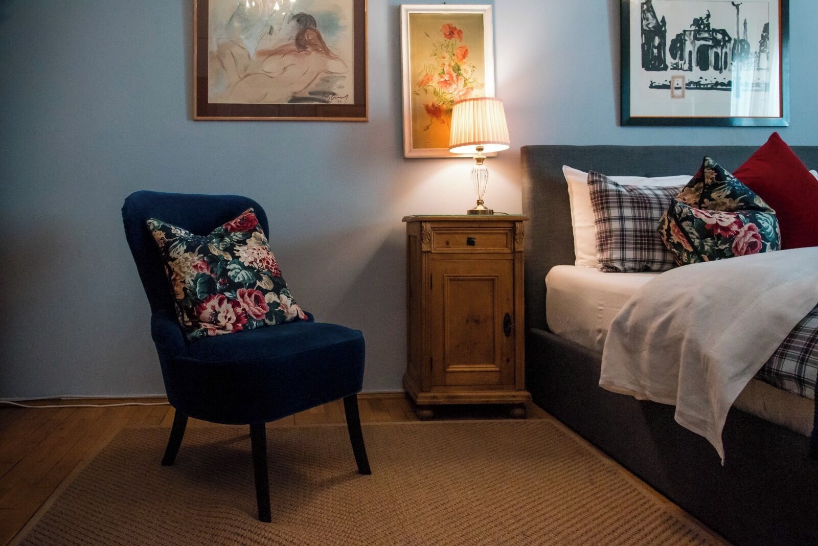 A room with a blue armchair with floral pillows, a wooden nightstand with a lamp, and a bed with plaid and floral pillows.
