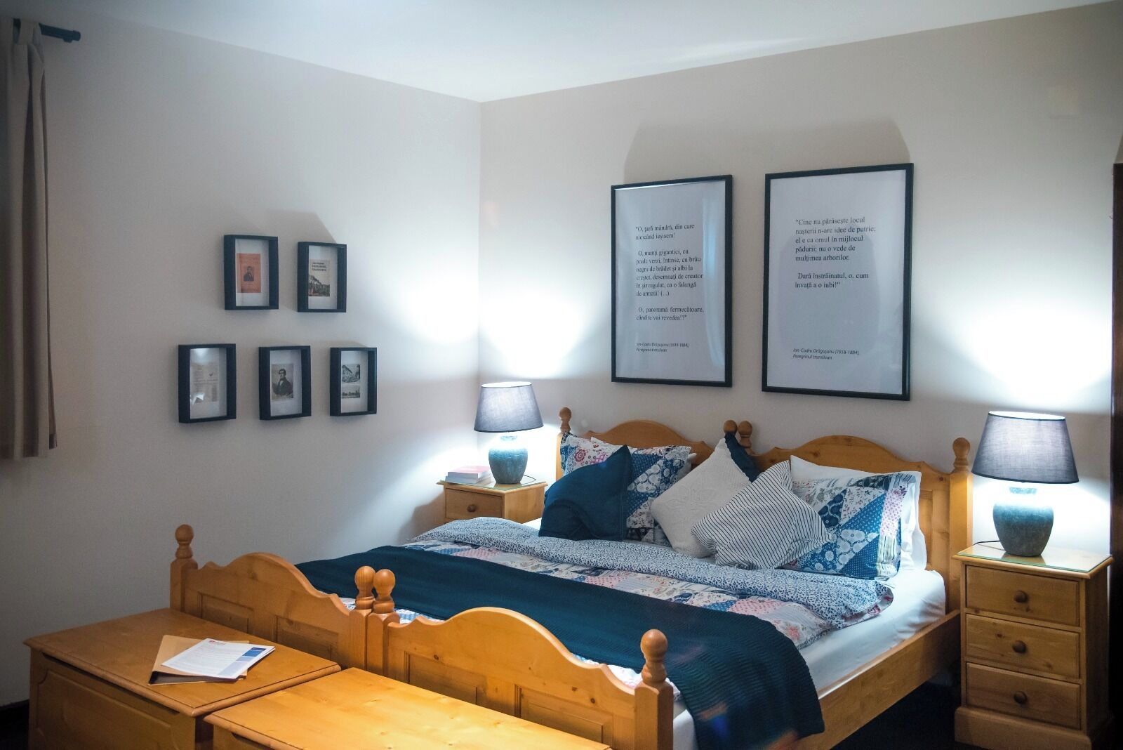 A neatly arranged bedroom with a wooden bed, a bedside table, a nightstand, and a small wooden desk. There are framed pictures on the wall and two framed quotes.