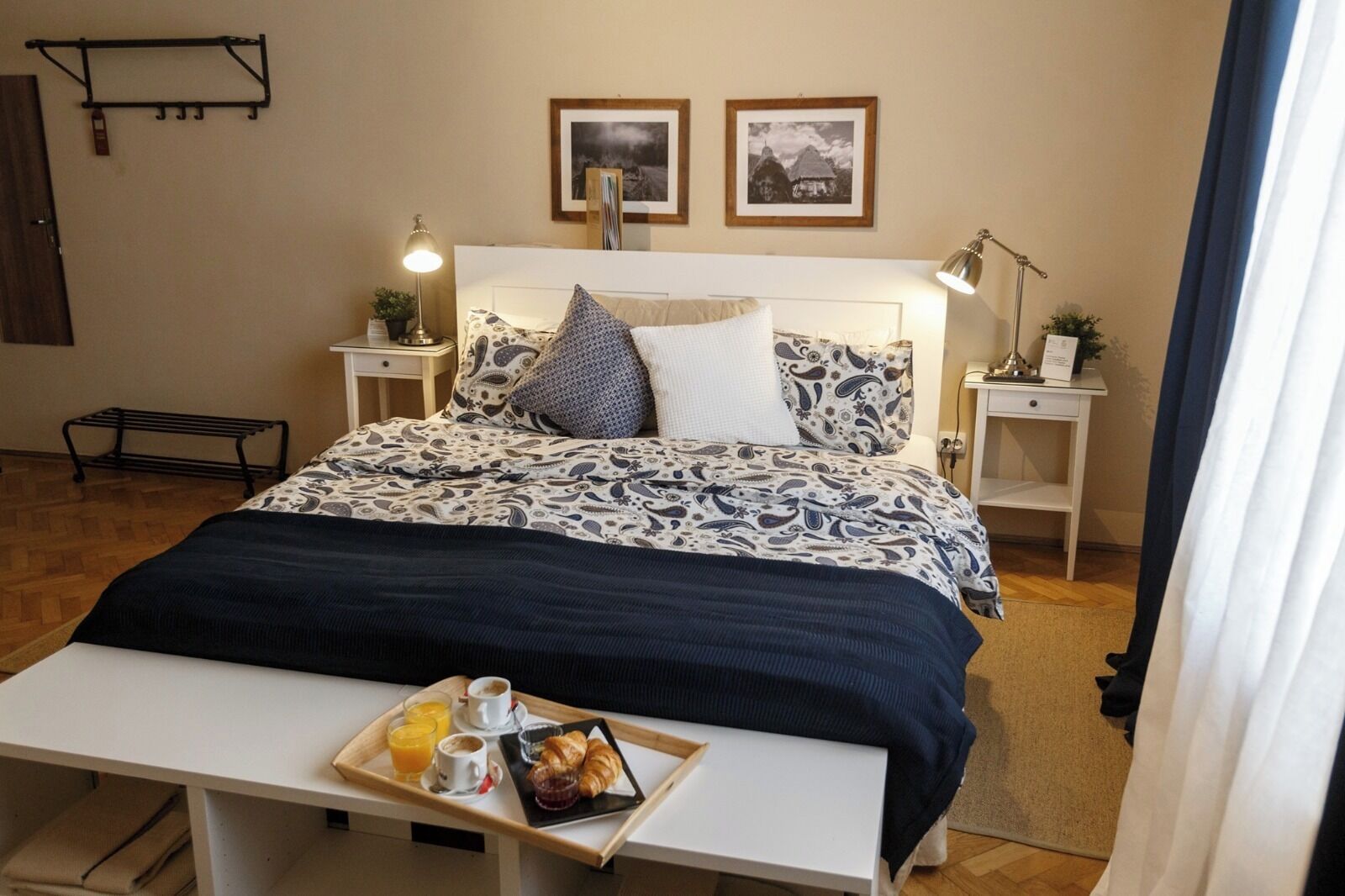 Una cama bien hecha con ropa de cama estampada, mesitas de noche con lámparas y una bandeja con alimentos para el desayuno sobre una mesa frente a la cama.