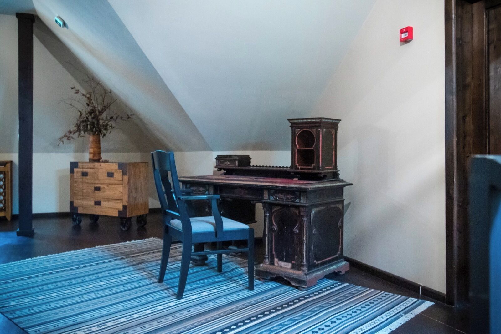Una habitación con techo inclinado, un escritorio de madera con lámpara antigua, una silla azul y un tapete estampado.