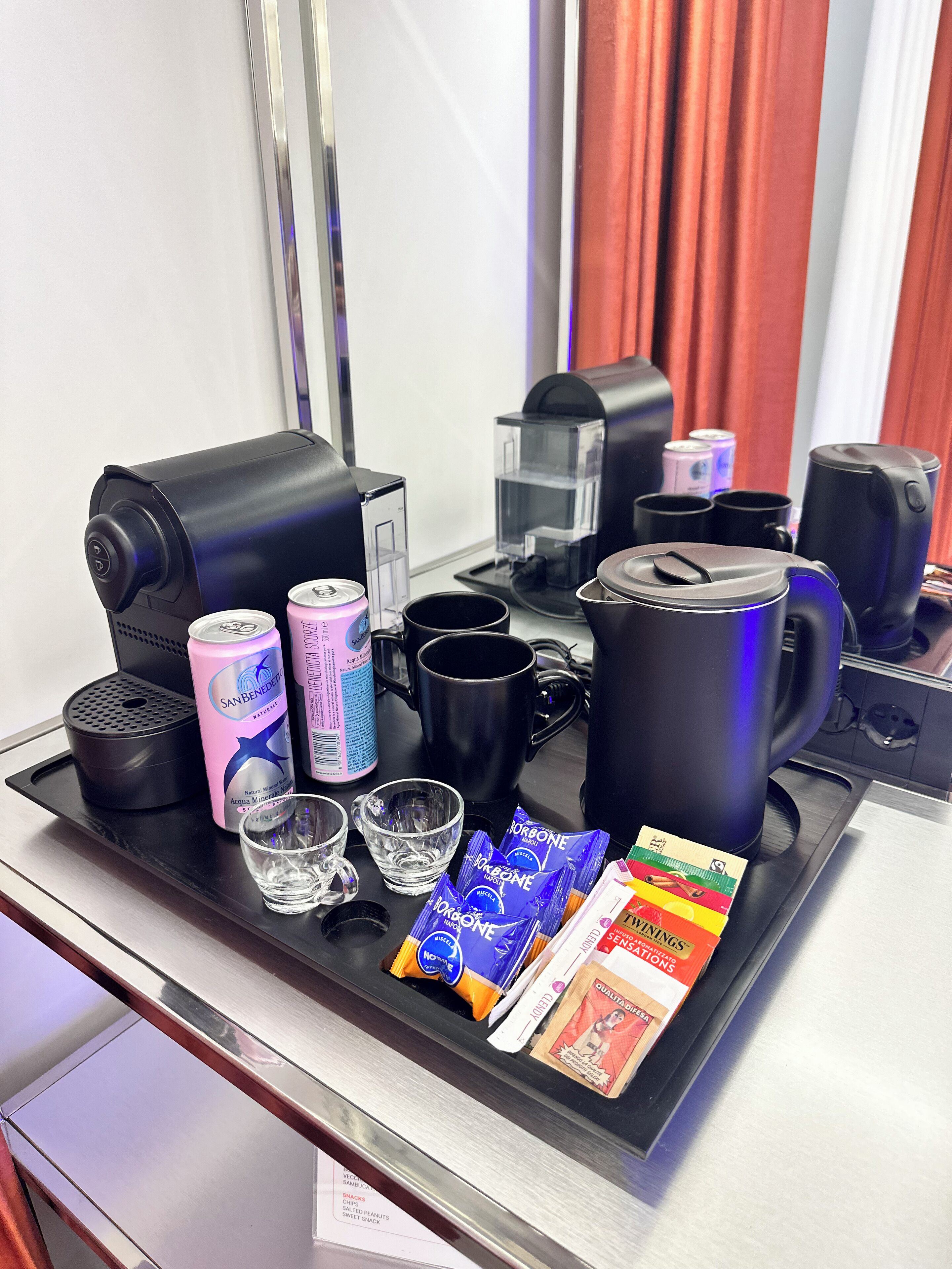 A breakfast tray with a coffee maker, cups, a water dispenser, a can of soda, a glass of water, and packaged snacks.