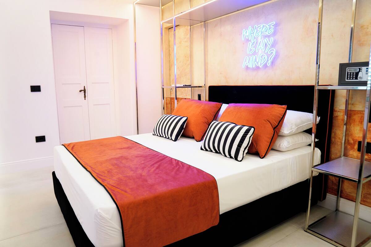 A neatly made bed with a red blanket, two orange pillows, and striped pillows.