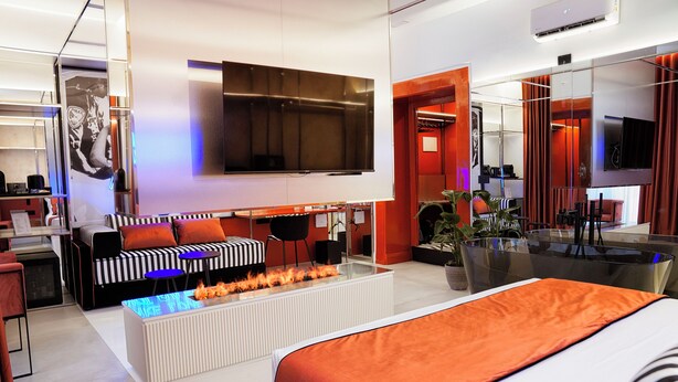 Una habitación de hotel moderna con una cama grande, un sofá, un televisor de pantalla plana, una mesa de centro de vidrio con chimenea integrada y un comedor con mesa y sillas de vidrio.