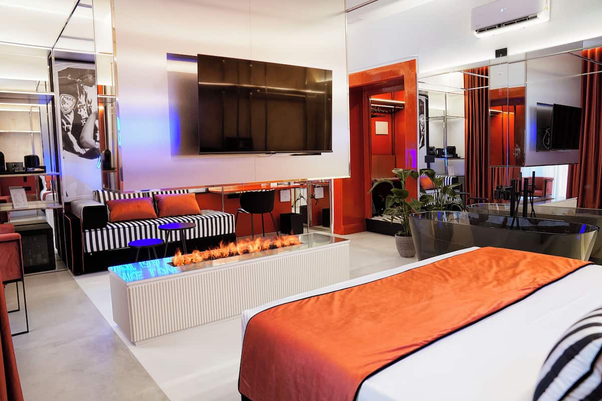 A modern hotel room with a large bed, a sofa, a flat-screen TV, a glass coffee table with a fire feature, and a dining area with a glass table and chairs.