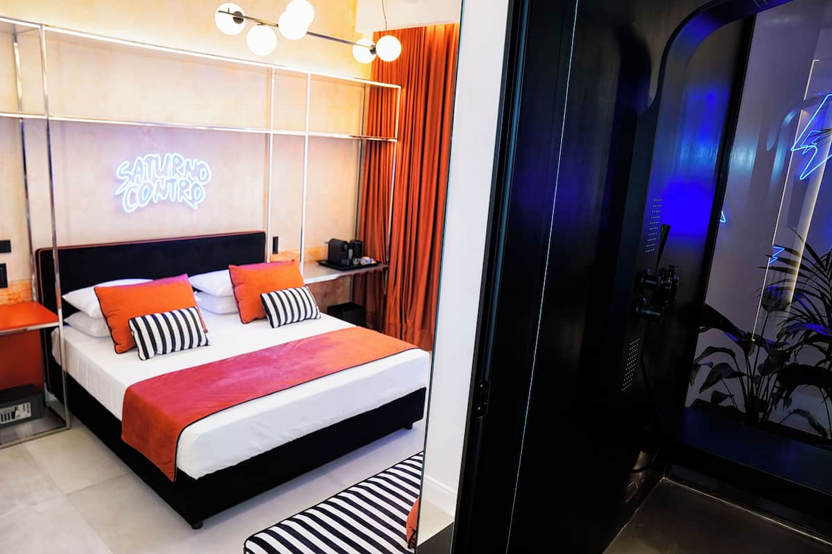 A modern hotel room with a bed, a neon sign, and a plant.