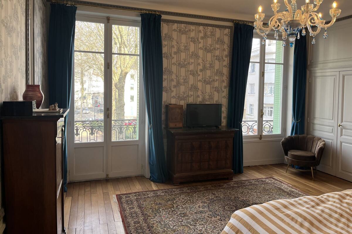 Una habitación con un aparador de madera, un televisor sobre un mueble bajo, una alfombra estampada, una cama con ropa de cama a rayas y una araña.
