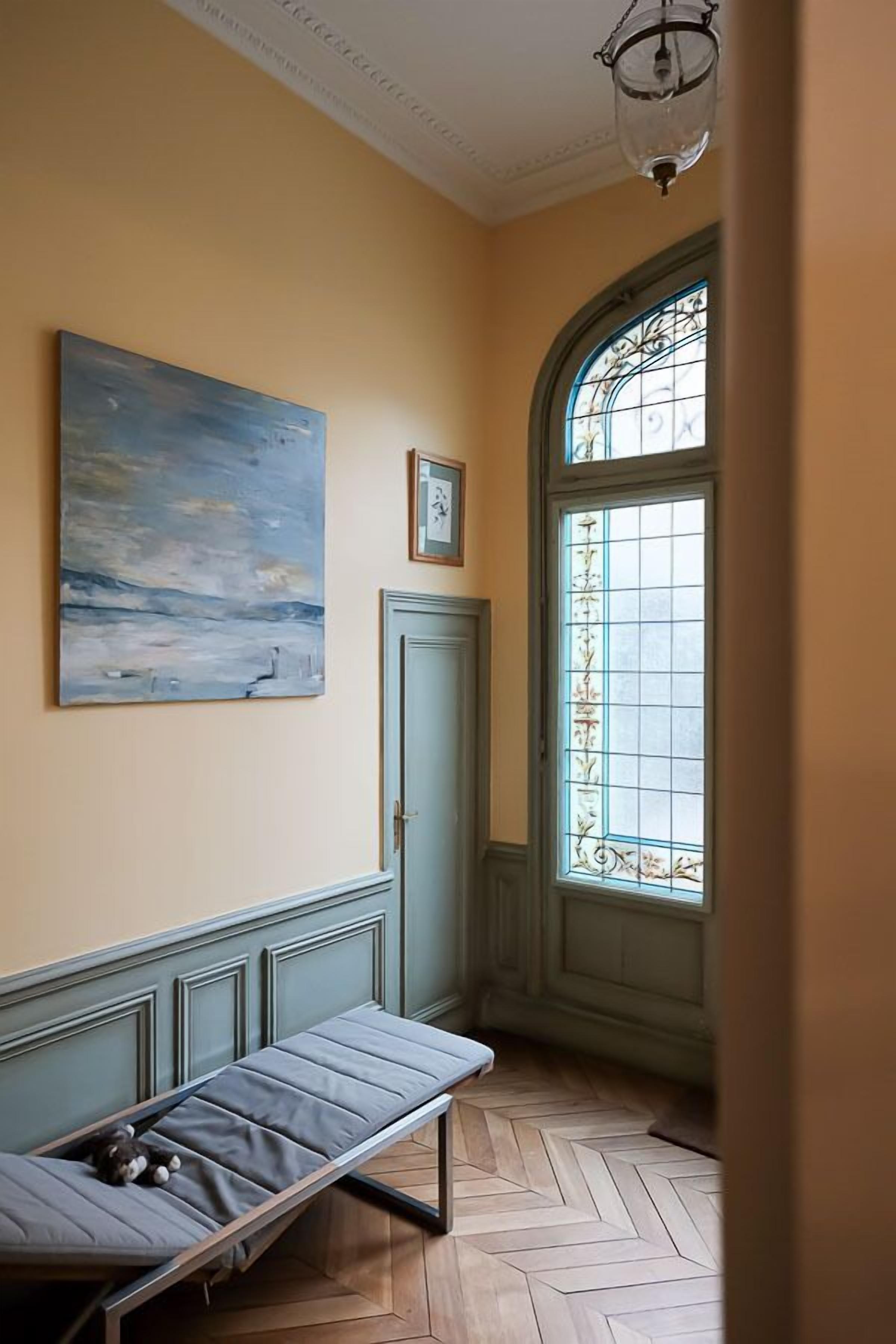 Un pasillo con un banco, un cuadro, una puerta y un ventanal con detalles decorativos.
