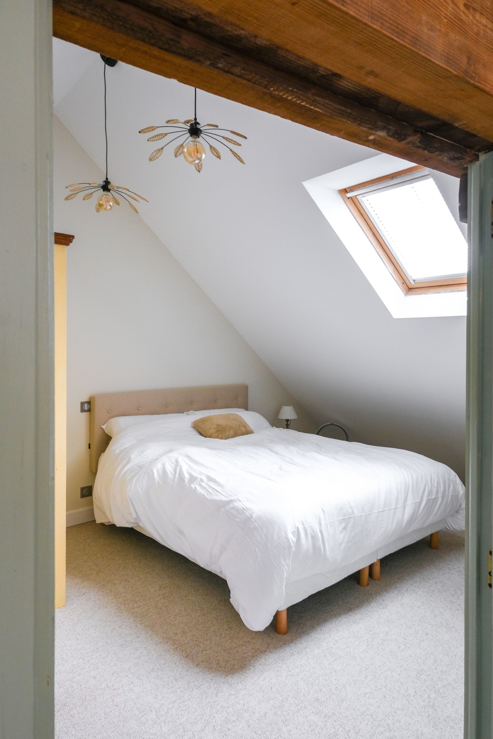 Ein Schlafzimmer mit schrägverdeckter Decke, einem Bett mit weißer Bettwäsche, einer einzelnen Nachttischlampe und einem Dachfenster.