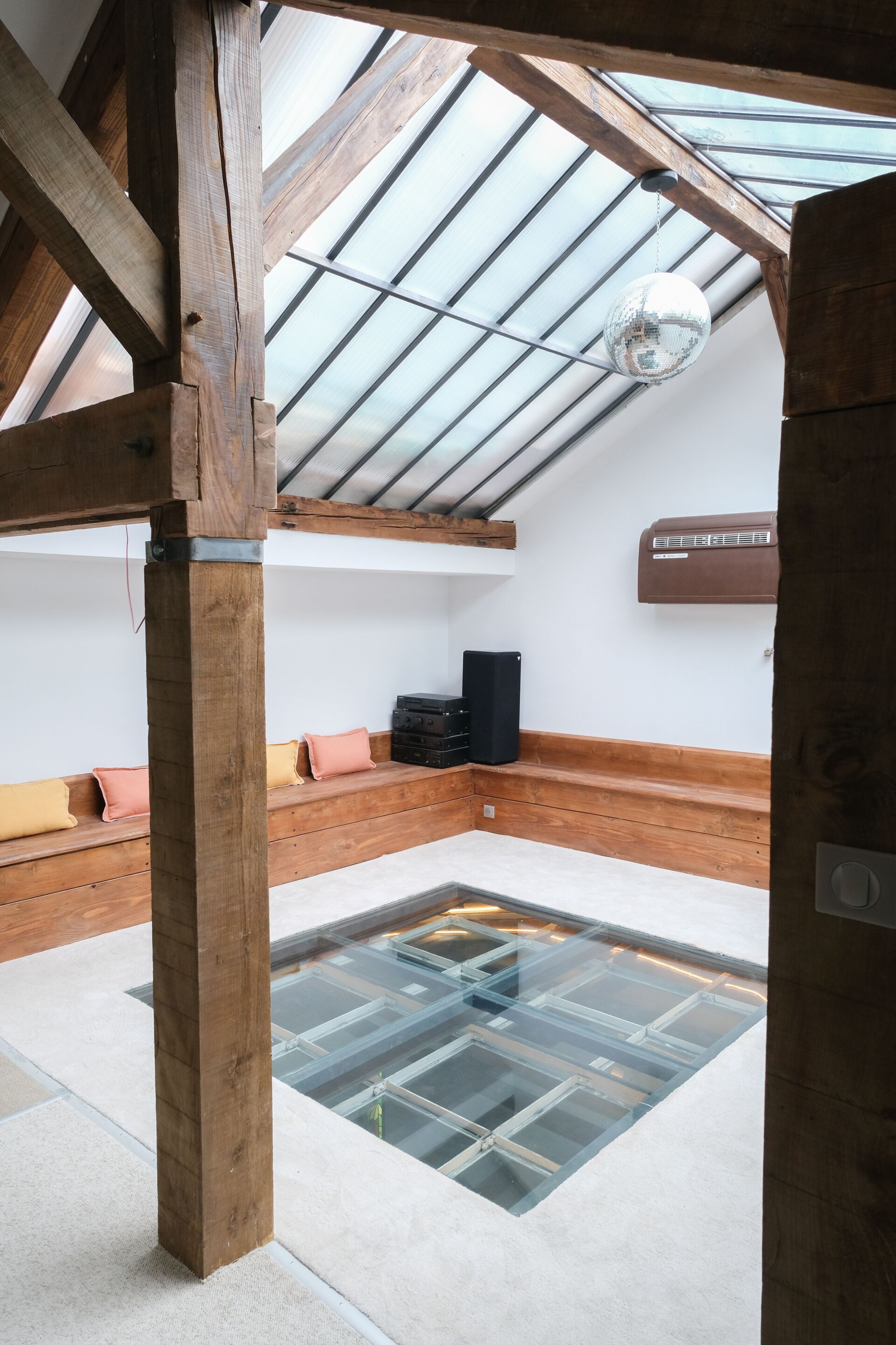 A room with a sloped ceiling, wooden beams, a skylight, a glass floor, and a black speaker on a wooden bench.