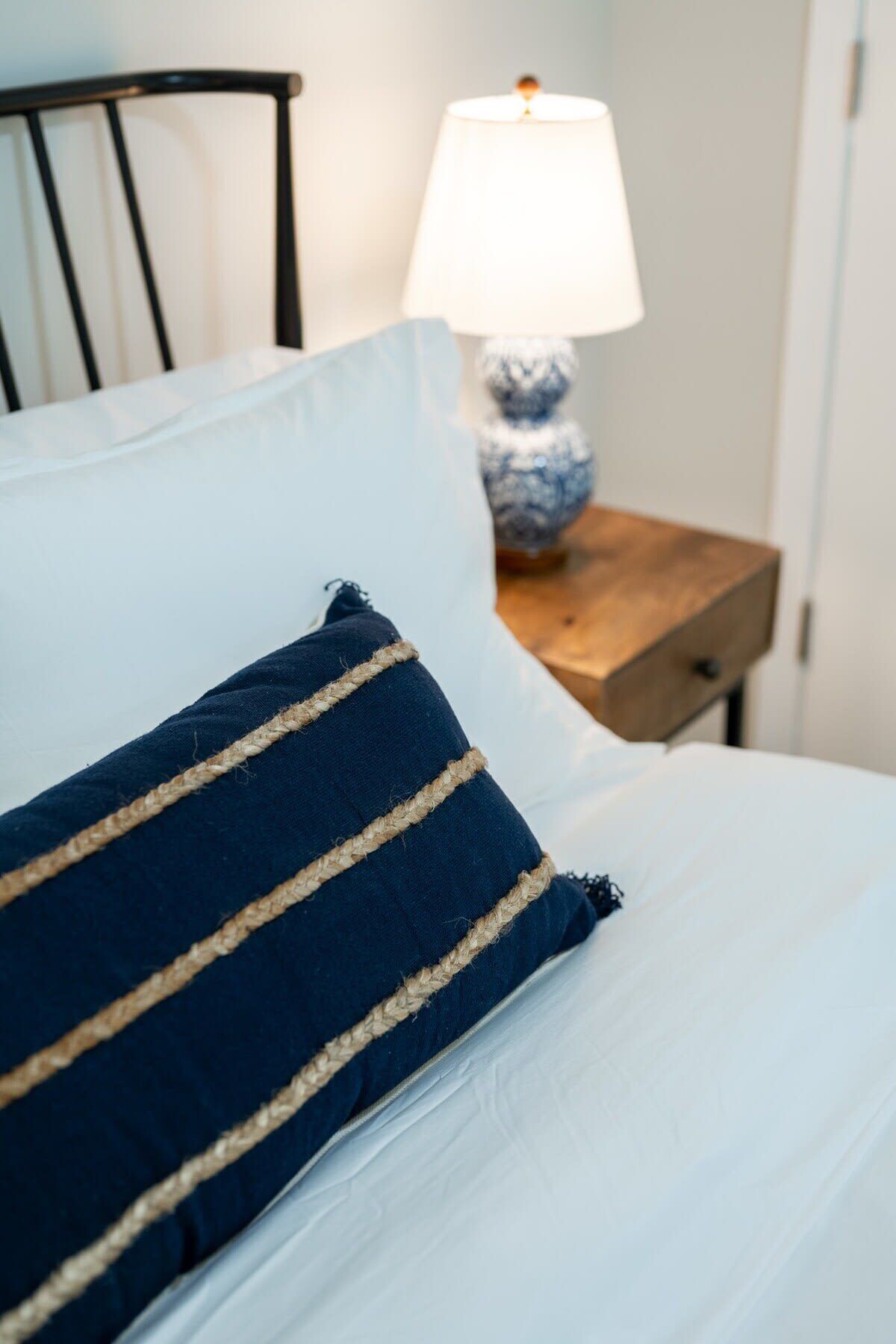 A bed with a white pillow and a dark blue pillow with rope trim. There is a wooden nightstand with a lamp and a decorative item beside the bed.