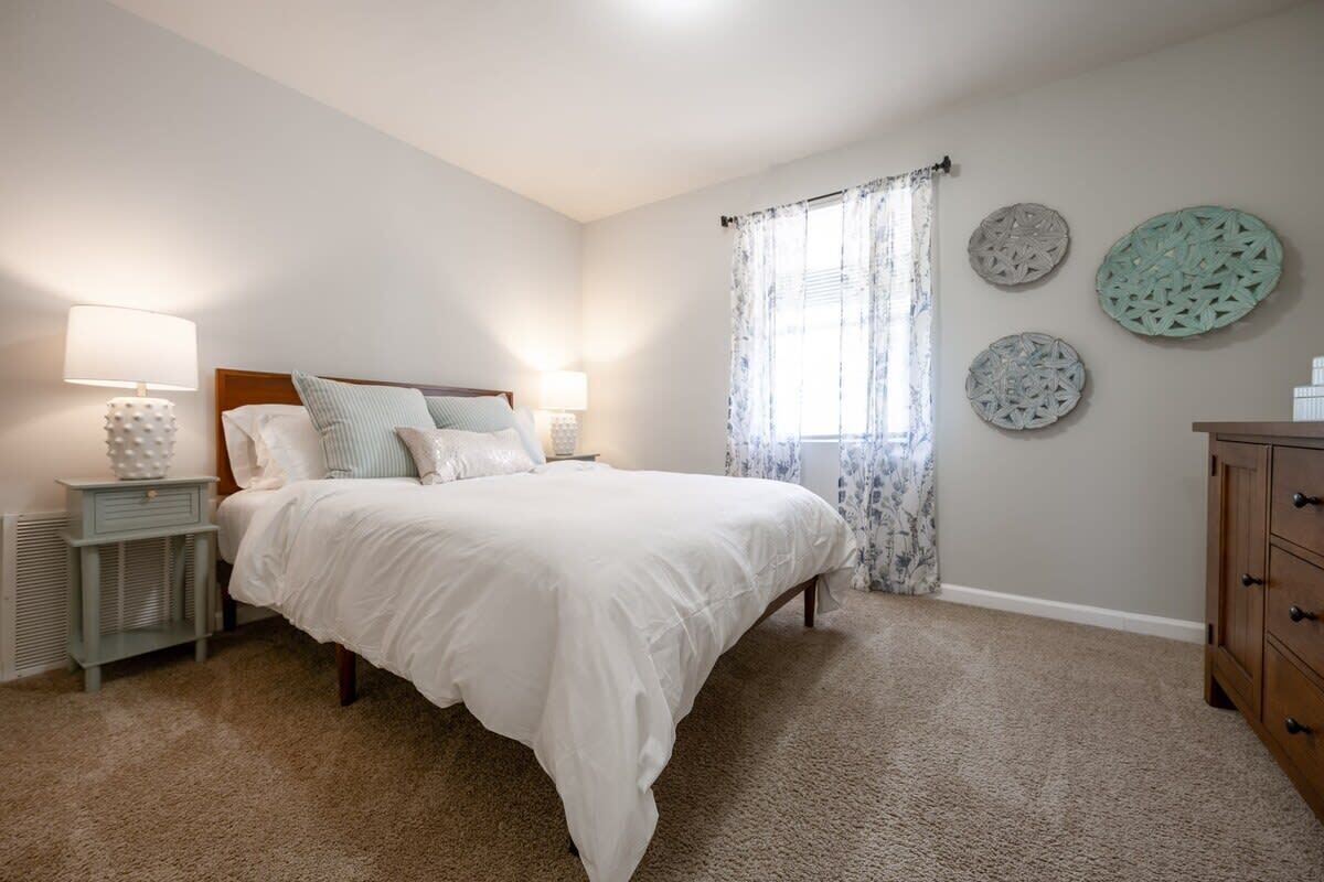 Quarto com cama, duas mesas de cabeceira, penteadeira e janela com cortinas.