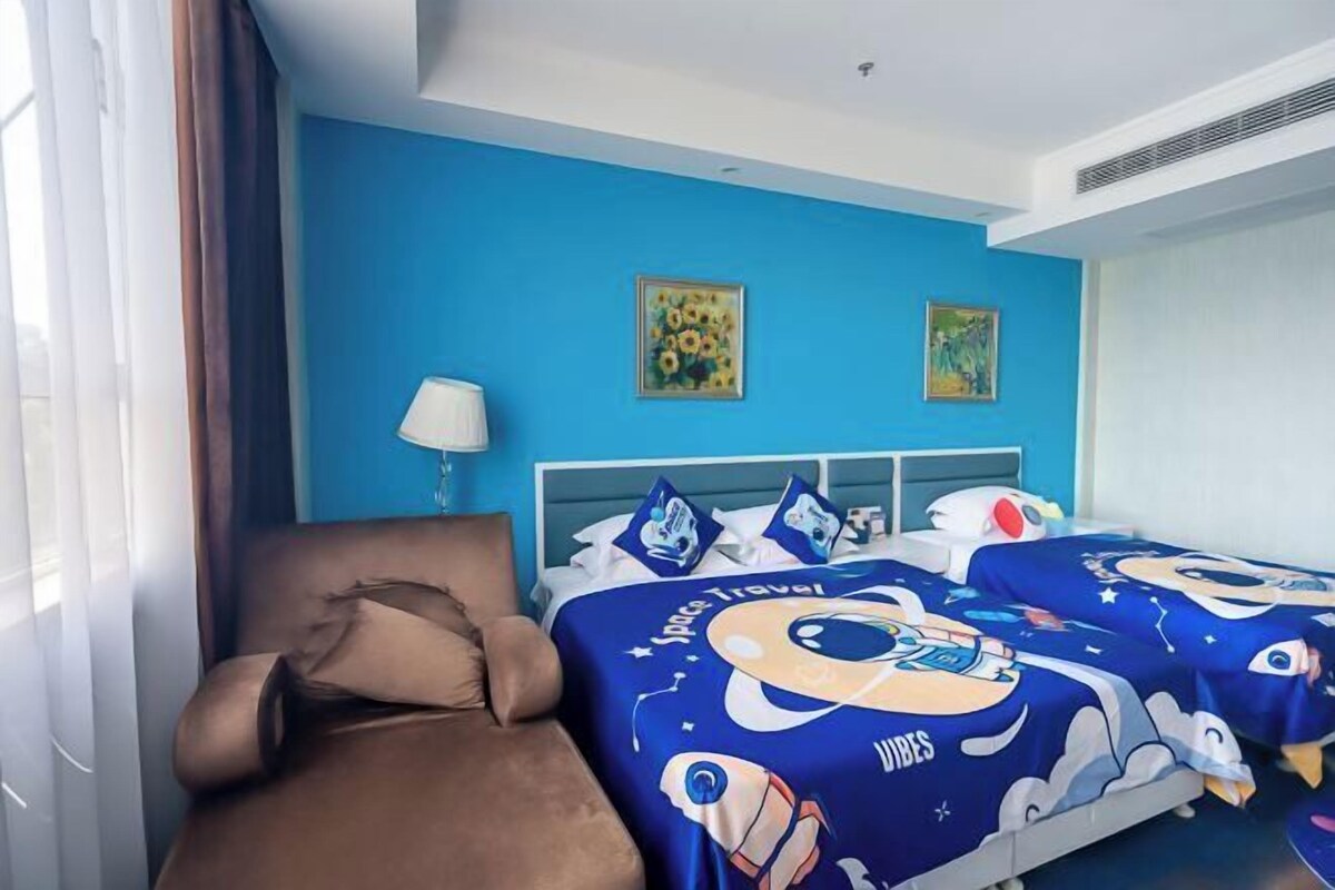 A hotel room with a blue wall, a brown sofa, two beds with space-themed bedding, and two paintings.