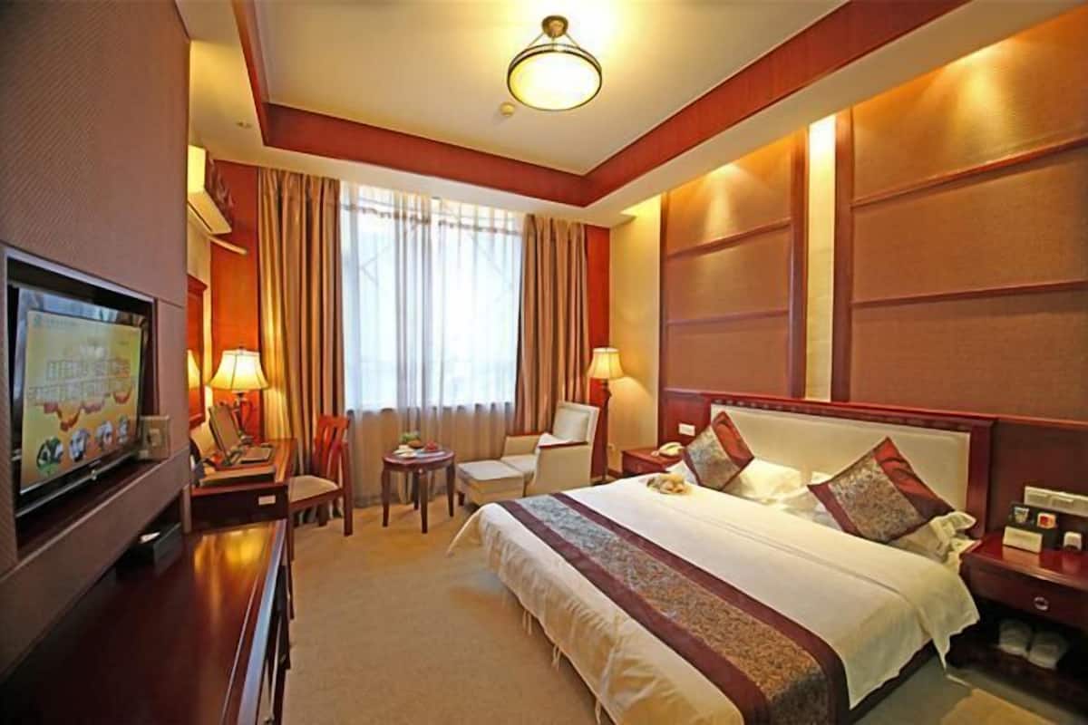A hotel room with a large bed, a desk with a TV, a chair, a small table, and a window with curtains.