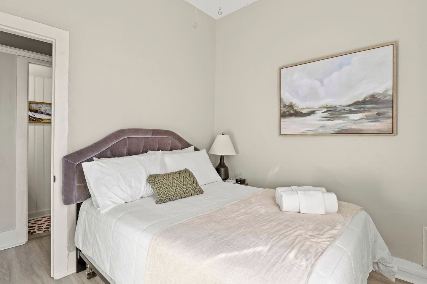 A neatly made bed with white linens and a decorative pillow, a bedside lamp, and a framed painting on the wall.