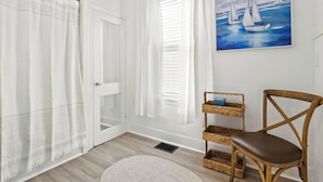 Una stanza con pavimento in legno, pareti bianche, una porta bianca, tende bianche, uno scaffale in vimini con un oggetto blu, una sedia di legno e un quadro con dei velieri.